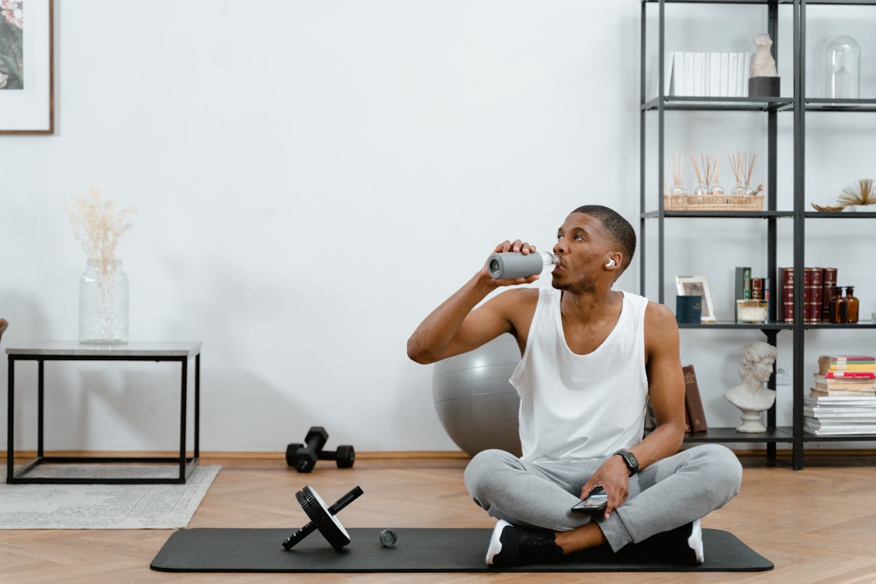 Adult man in active wear resting on yoga mat, drinking water post-workout indoors.